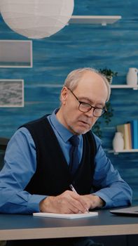 Multitasking retired man reading on laptop and writing on notebook