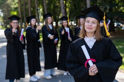 Group of happy students in graduation gowns outdoors. A young girl with a diploma in her hands in the foreground.