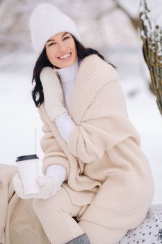A beautiful girl with a beige cardigan and a white hat enjoying drinking tea in a snowy winter forest near a lake
