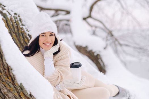 A beautiful girl with a beige cardigan and a white hat enjoying drinking tea in a snowy winter forest near a lake