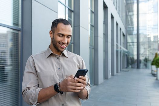 hispanic man outside modern office building using smartphone, businessman in shirt typing message and browsing online pages