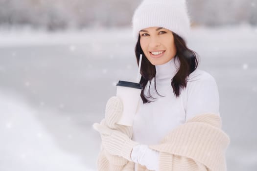 A beautiful girl in a beige cardigan and a white hat with a glass of tea enjoys a snowy embankment by the lake