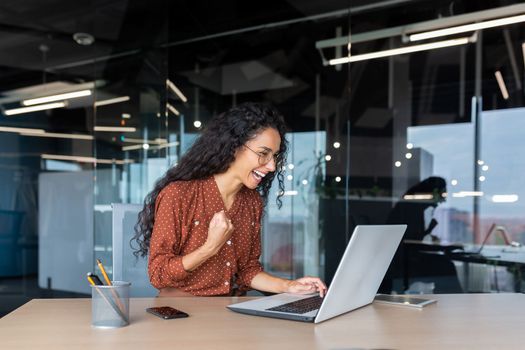 Hispanic businesswoman celebrating victory and successful achievement, office worker received online news and happy smiling and looking at laptop screen holding hand up triumph gesture