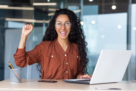 Portrait of a successful hispanic businesswoman working inside a modern office building smiling and looking at the camera, holding her hand up in a gesture of success and triumph