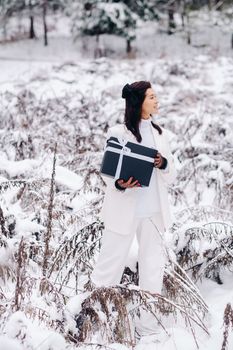 A stylish woman with a white suit with a New Year's gift in her hands in a winter forest. A girl in nature in a snowy forest with a gift box