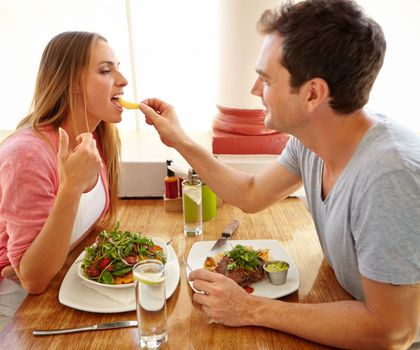 Isnt this better than a salad. A handsome young man giving his girlfriend a taste of his food while they have lunch together in a restaurant.