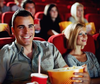 Ive been looking forward to this all week. A portrait of a happy young man holding a refreshment and popcorn in a cinema while with his girlfriend.