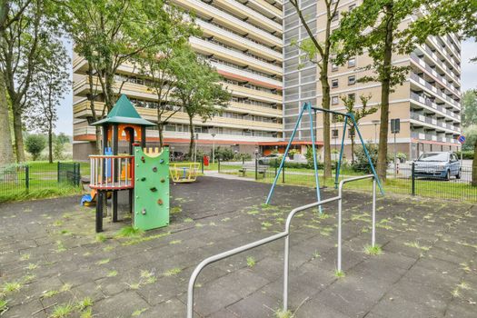 an empty playground in front of a multi - storey apartment building, with children's play equipment and cars parked on the pavement