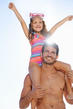 Happy to be on holiday. A happy young girl wearing a swimsuit sitting on her fathers shoulders with her hands in the air.