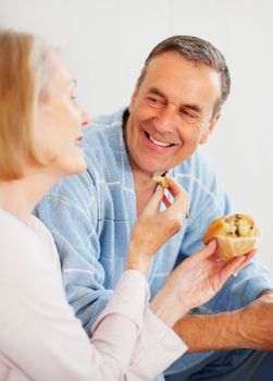 Woman feeding muffin to husband against colored background. Portrait of a loving mature woman feeding muffin to husband against colored background.