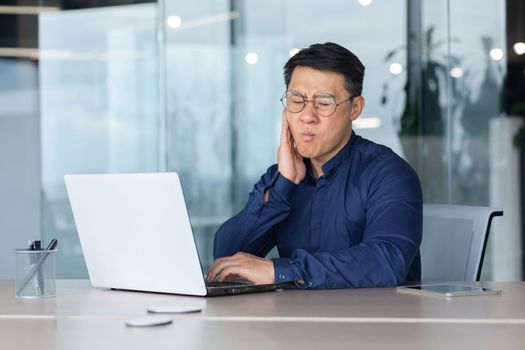 A young Asian man works in the office at a desk with a laptop. He holds his cheek. He feels a strong toothache, he needs the help of a dentist.
