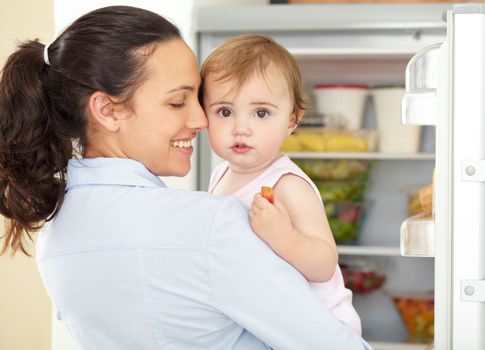 She can be fussy sometimes. a cute baby being held by its caring mom in front of an open fridge.