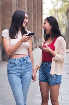 two young women sharing confidences and having fun with their cell phones while walking through the city holding hands, concept of friendship and love between people of the same sex