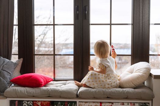 Girl 2 years old sits on the windowsill. A little blonde girl in a white dress on the windowsill looks out the window and holds a candy in her hand.