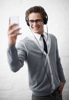 His phone doubles up as a mp3 player. A young nerdy guy looking seriously at his phone while wearing headphones.