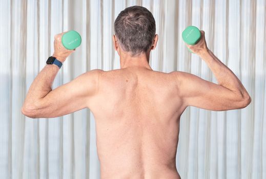 Senior man shirtless and exercising in bedroom with dumbbell
