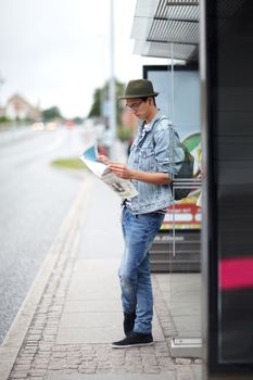 Picking his route. Hip young guy standing at a bus stop while looking at a map.