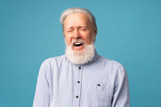 Retired old man with white hair and beard open mouth laughter excited over blue color background