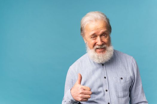 Cheerful senior bearded old man show fingers thumb up select over blue color background with copy space
