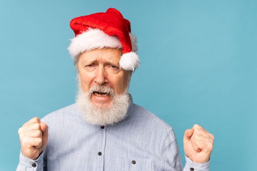 Happy confident cool old bearded Santa Claus winner raising fists celebrating triumph and success over blue background with copy space