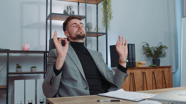 Businessman working on laptop, meditating, doing yoga breathing exercise in lotus position at office