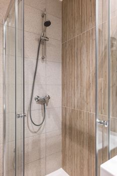 Shower area with marble tiles on the walls. On the wall is a faucet with a long hose and a shower head. Area is fenced with glass doors to protect against splashing water.