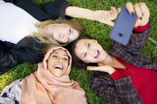 Multiracial girlfriends laughing while lying on lawn and taking selfie