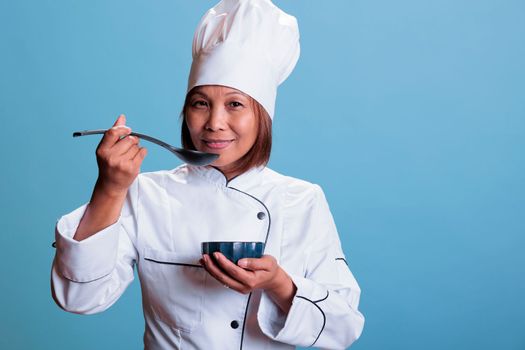 Cheerful cook with restaurant uniform holding bowl with culinary meal while tasting food