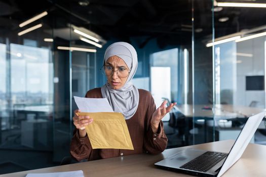 Upset business woman in hijab working inside office, woman received letter envelope mail with bad news, office worker upset by notification working at workplace with laptop