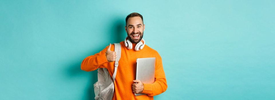 Happy man with backpack and headphones, holding laptop and smiling, showing thumb-up in approval, standing over turquoise background