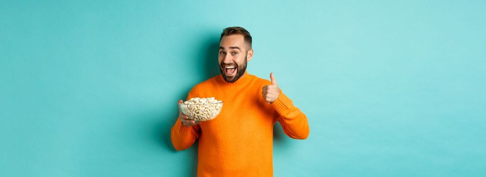 Satisfied young man, showing thumb-up and smiling, eating popcorn and watching good movie or tv, standing over blue background