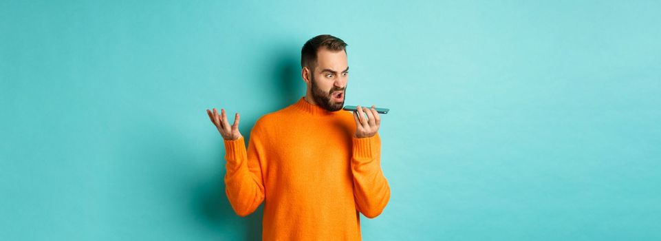 Angry man arguing on speakerphone, record voice message with mad face, standing over light blue background in orange sweater
