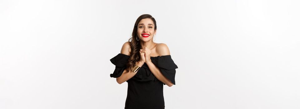 Fashion and beauty. Pretty woman in black dress asking for help, showing thank you gesture, looking grateful at camera, standing over white background