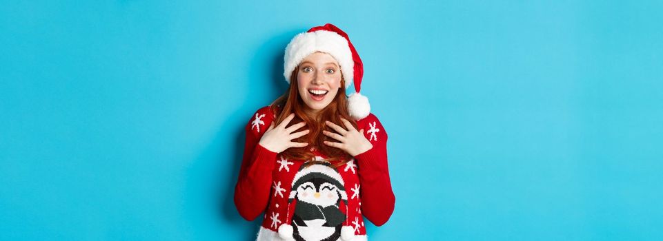 Happy holidays and Christmas concept. Surprised redhead girl receiving unexpected gift, gasping amazed and staring with plesant disbelief, standing in santa hat against blue background