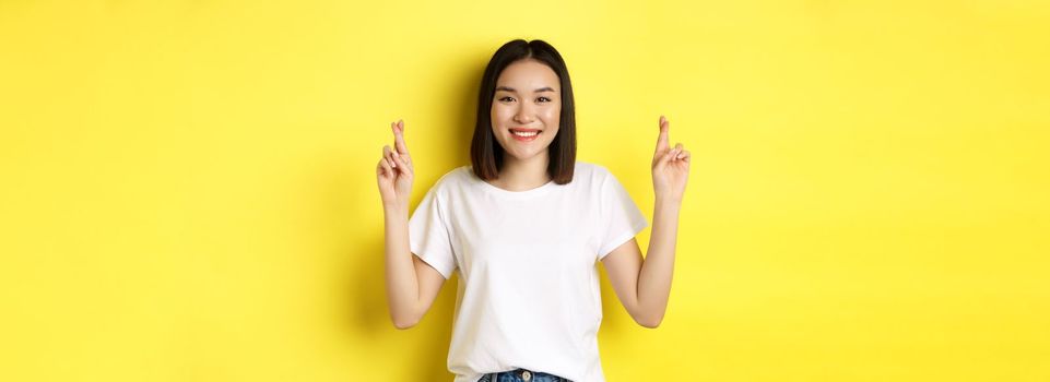 Hopeful asian girl making wish, cross fingers for good luck and praying with eyes closed, standing over yellow background