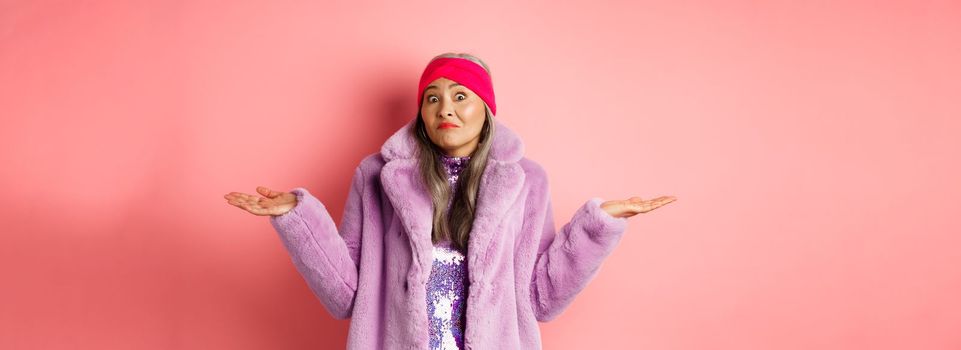 Fashionable and funky asian old woman shrugging shoulders, looking confused and clueless, standing in stylish purple coat and headband, pink background