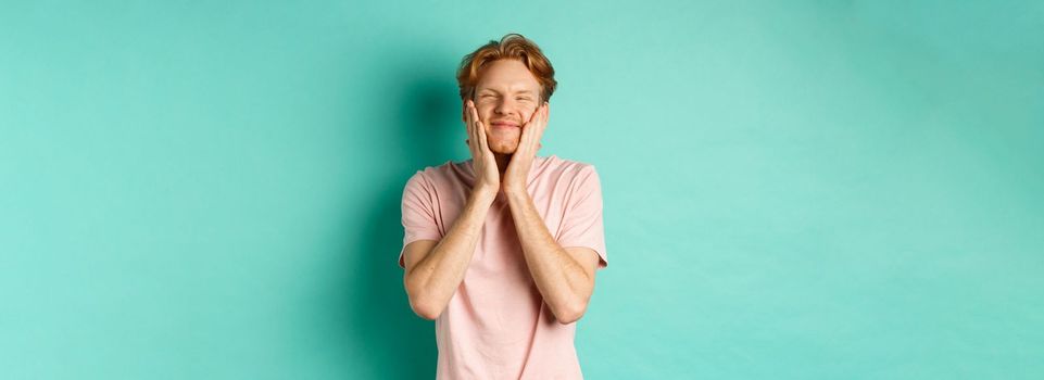 Image of cute and silly young man with red hair, feeling satisfaction while touching his face, smiling happy, standing over turquoise background
