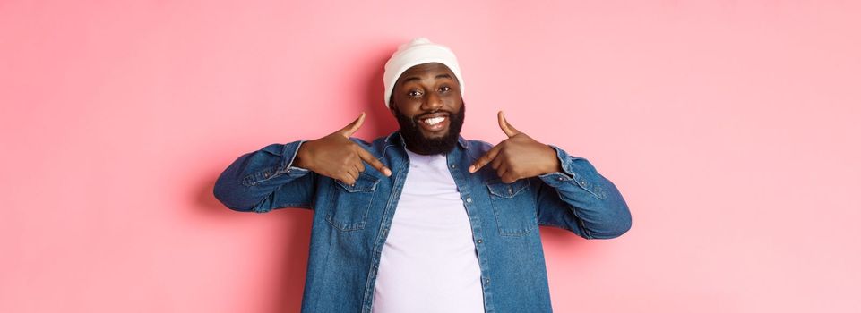Happy young african-american hipster guy pointing at himself t-shirt, smiling pleased, standing over pink background