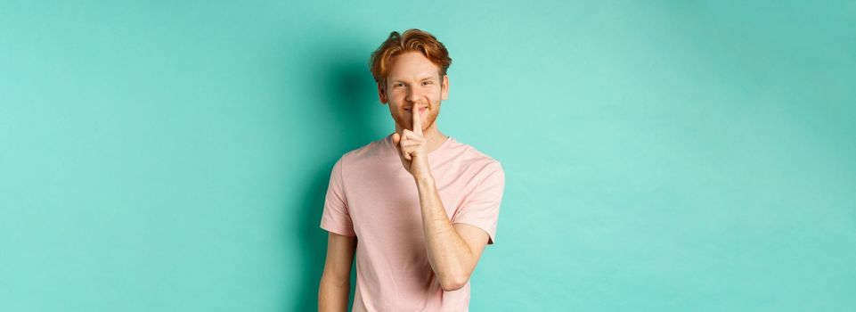 Smiling young man with red hair and beard sharing a secret, showing taboo gesture and grinning, shushing you to be quiet, standing over turquoise background