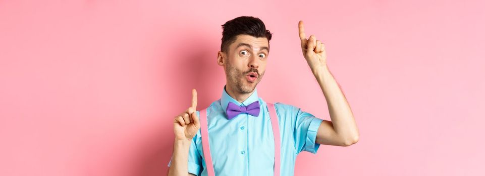 Cheerful funny man dancing in bow-tie and suspenders, pointing fingers up and looking upbeat, standing over pink background