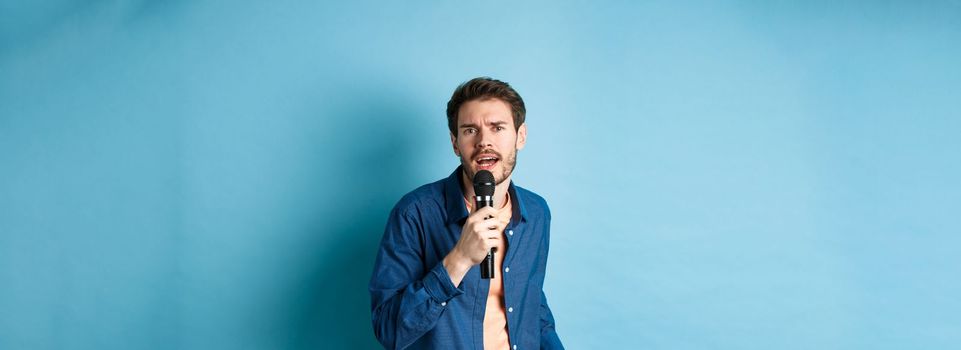 Passionate singer looking at camera, singing in microphone, playing karaoke on blue background