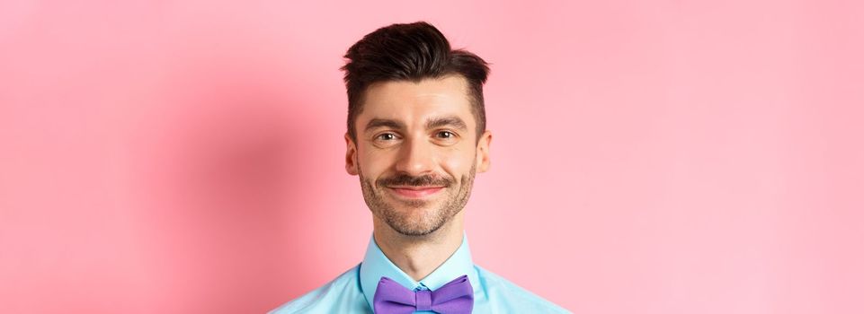 Close-up of smiling hansome man with moustache, wearing bow-tie and shirt, standing cheerful on pink background