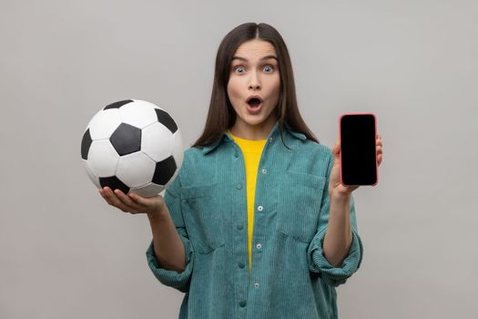 Surprised woman holding and showing blank display smartphone and football ball, betting on soccer.