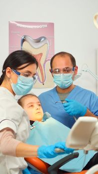 Little girl patient sitting in stomatological chair in dental office, visiting doctor for oral problems. Woman dentist with protective gloves and mask removes tooth decay drilling teeth during surgery