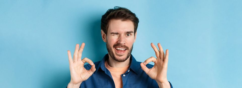 Close up of handsome confident guy winking, showing okay gestures, guarantee quality, recommending something good, standing on blue background