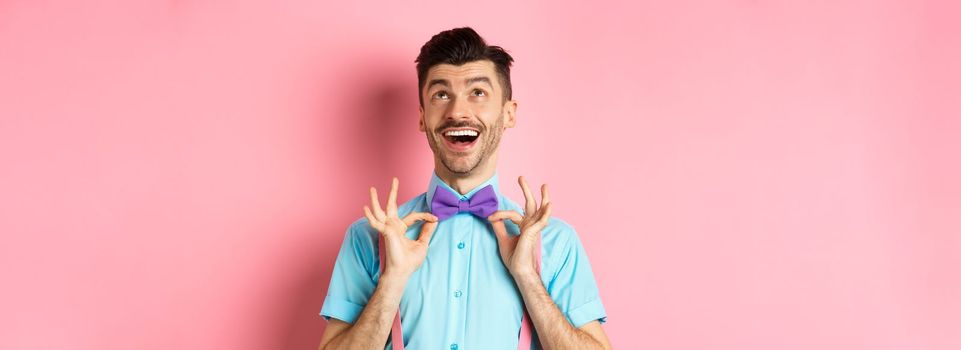 Happy young man with moustache, touching his bow-tie and laughing, looking up at logo, standing on pink background