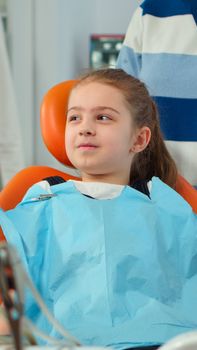 Close up of kid patient with toothache wearing dental bib talking with dentist before intervetion showing to affected mass. Girl sitting on stomatological chair while nurse preparing sterilized tools.