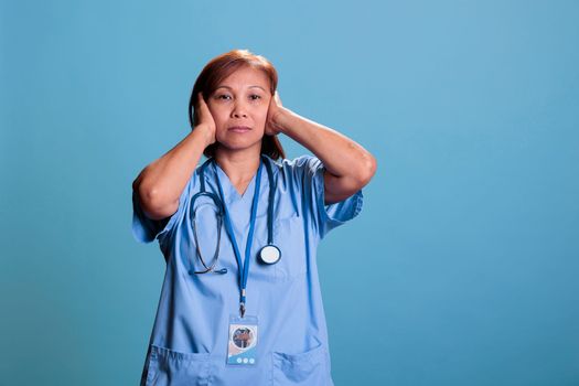 Asian quiet nurse with stethoscope covering ears having serious expression
