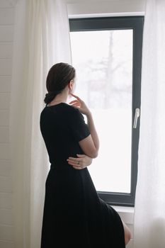 Silhouette of young woman standing by the window. Emotional portrait of a beautiful lonely girl with dark hair indoors