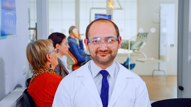 Portrait of stomatologist smiling at camera being in dental office while patients waiting him in background. Dentistry doctor looking on webcam sitting on chair in stomatological clinic.